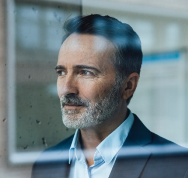 Man in a suit with a gray beard looking out of a window, partial reflection visible in the glass