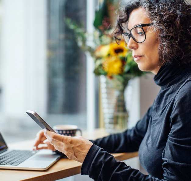 Frau, am Laptop sitzend mit Smartphone in der Hand