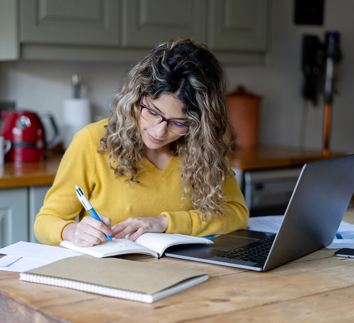 Image of a woman who is working on her laptop and going through notes in her notebook.